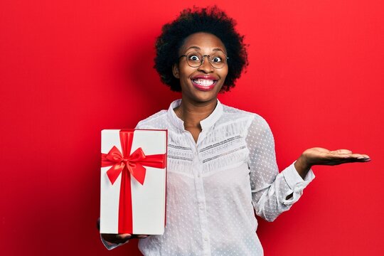 Young african american woman holding gift celebrating achievement with happy smile and winner expression with raised hand