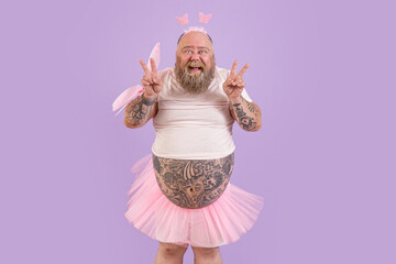 Excited middle aged man with overweight in carnival fairy suit with pink wings shows peace gestures...