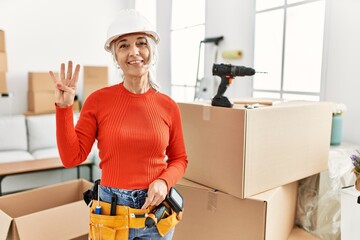 Middle age grey-haired woman wearing hardhat standing at new home showing and pointing up with fingers number four while smiling confident and happy.