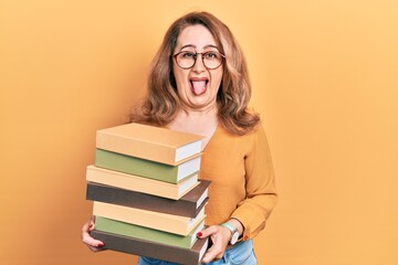 Middle age caucasian woman holding a pile of books sticking tongue out happy with funny expression.