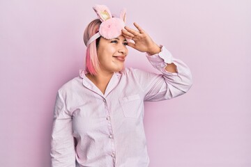 Hispanic woman with pink hair wearing sleep mask and pajama very happy and smiling looking far away with hand over head. searching concept.