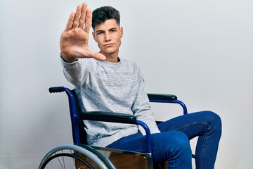 Young hispanic man sitting on wheelchair doing stop sing with palm of the hand. warning expression...