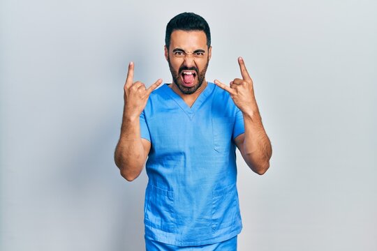 Handsome hispanic man with beard wearing blue male nurse uniform shouting with crazy expression doing rock symbol with hands up. music star. heavy concept.