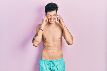 Fototapeta premium Young hispanic man wearing swimwear shirtless covering ears with fingers with annoyed expression for the noise of loud music. deaf concept.