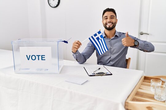 Young handsome man with beard at political campaign election holding greece flag smiling happy and positive, thumb up doing excellent and approval sign