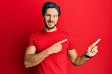 Young hispanic man wearing casual red t shirt smiling and looking at the camera pointing with two hands and fingers to the side.