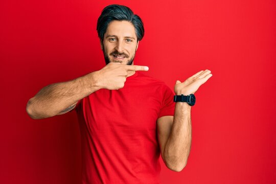 Young hispanic man wearing casual red t shirt amazed and smiling to the camera while presenting with hand and pointing with finger.