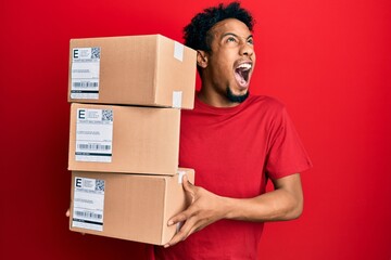 Young african american man with beard holding delivery packages angry and mad screaming frustrated and furious, shouting with anger looking up.