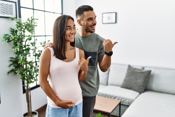 Fototapeta na wymiar Young interracial couple expecting a baby, touching pregnant belly smiling with happy face looking and pointing to the side with thumb up.