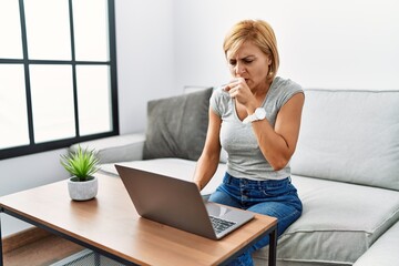 Middle age blonde woman using laptop at home feeling unwell and coughing as symptom for cold or bronchitis. health care concept.