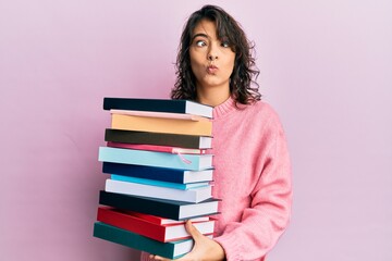 Young hispanic woman holding a pile of books making fish face with mouth and squinting eyes, crazy and comical.