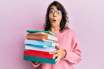 Young hispanic woman holding a pile of books scared and amazed with open mouth for surprise, disbelief face