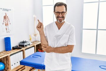 Middle age man with beard working at pain recovery clinic with a big smile on face, pointing with hand and finger to the side looking at the camera.