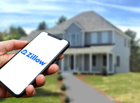 Zillow logo is displayed on a smartphone in the hand of a person, in the background a house, Online real estate marketplace, real estate, house purchase, 2022