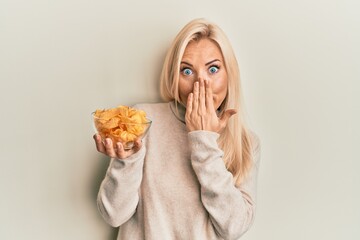 Young blonde woman holding bowl with potato chip covering mouth with hand, shocked and afraid for...