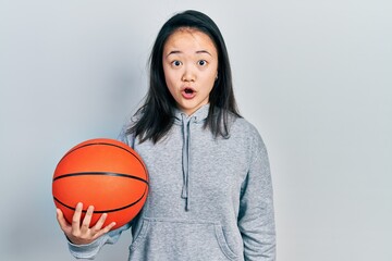 Young chinese girl holding basketball ball scared and amazed with open mouth for surprise, disbelief face