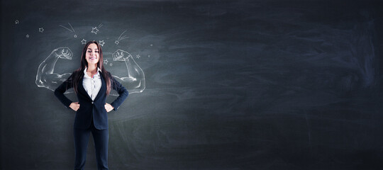 Attractive young european businesswoman standing on blackboard wall background with drawn muscle...
