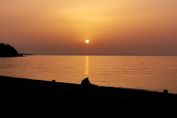 Silhouette of two people sitting on the beach during the sunrise. Sun rises over the Mediterranean Sea on a lovely summer morning. Selective focus.