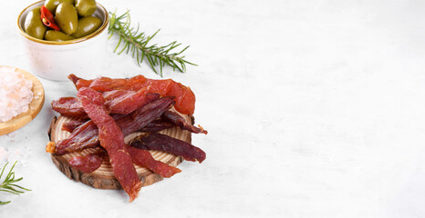 Jerky meat strips with spices, green olives, rosemary on wooden plate on a grey stone background. Tipical Italian food coppiette is a spiced pork meat strips. Snacks for beer, top view, copy space