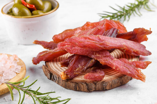 Jerky meat strips with spices, green olives, rosemary on wooden plate on a grey stone background. Tipical Italian food coppiette is a spiced pork meat strips. Snacks for beer, close up