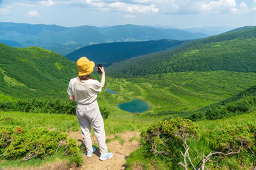 Fototapeta na wymiar A traveler in a hat and a backpack is resting in nature. A tourist girl photographs the panorama of the mountains and the lake. Mountain beautiful landscape