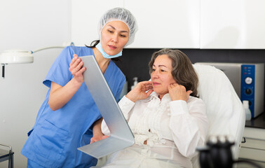 Female cosmetologist giving consultation to mature woman patient lying on couch in aesthetic clinic