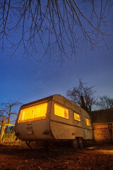 Old white caravan trailer with yellow illumination in backyard at stary blue night