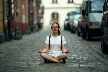 A teenage girl is sitting in the lotus position in the middle of the city pavement.