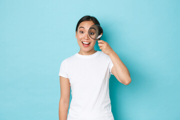 Cheerful smiling beautiful asian girl searching for someone, hiring to company, looking through magnifying glass with amused upbeat expression, standing over light blue background - obrazy, fototapety, plakaty