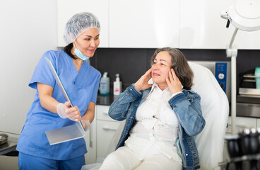 Smiling aged woman looking at mirror while professional cosmetician showing her results of hardware facial procedure in medical esthetic office.