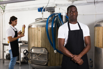 Serious man brewer standing in beer factory during work, man on background