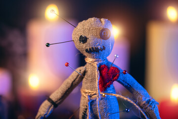 Voodoo doll studded with needles with pierced red rag heart against background of burning candles....