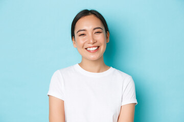 Close-up of cheerful carefree asian girl in white t-shirt smiling broadly. Pleasant korean female looking friendly at camera, enjoying nice conversation, talking with someone, light blue background - Powered by Adobe