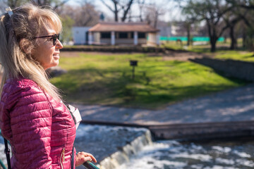 Side view of a woman standing in the park with a pond and green grass behind looking away