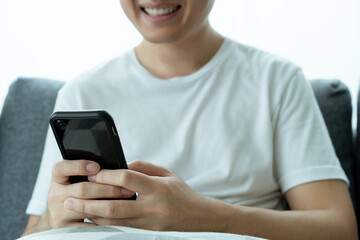 Asian young man in white t-shirt using a touchscreen smartphone while staying in the living room close up. Man browsing the internet and social media on his smartphone.