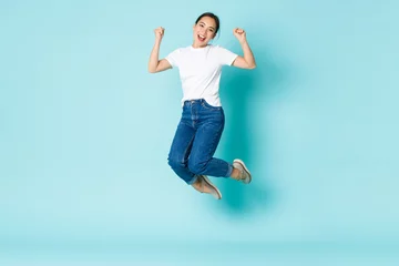Foto op Plexiglas Fashion, beauty and lifestyle concept. Cheerful triumphing, attractive asian girl jumping from happiness and joy, winning competition, celebrating victory over light blue background © Mix and Match Studio