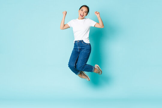 Fashion, beauty and lifestyle concept. Cheerful triumphing, attractive asian girl jumping from happiness and joy, winning competition, celebrating victory over light blue background