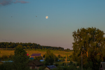 a full moon hangs over a small village on a warm summer morning