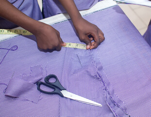 Female african tailor's hands measuring a fabric with tape measure during a fashion designing training session or class, scissors on the table