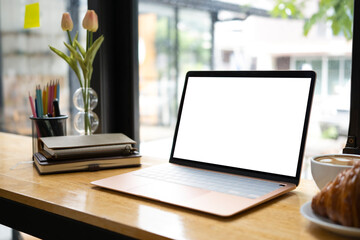 Computer laptop with empty display, books and coffee cup on wooden table in cafe.