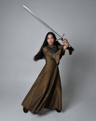 Full length  portrait of beautiful young asian woman with long hair wearing medieval fantasy gown costume. Graceful standing posing holding a sword,  isolated on studio background.