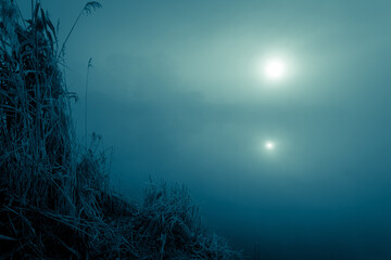 Night mystical scenery. Full moon over the foggy river and its reflection in the still water, frosty reeds on the bank on the foreground.