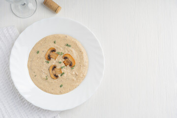 Top view of creamy mushroom soup puree made of blended champignons, potatoes and dairy cream decorated with fried slices and thyme served with glass of wine on plate on white wooden table. Copy space
