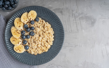 Top view of oatmeal porridge made of boiled oats decorated with fresh blueberries and sliced banana served on blue gray plate on textile on concrete background for breakfast. Image with copy space