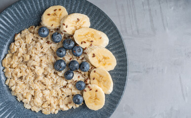 Top close view of healthy oatmeal porridge made of cooked oats decorated with sweet blueberries, sliced banana and flax seeds served on blue gray plate on concrete background for breakfast. copy space