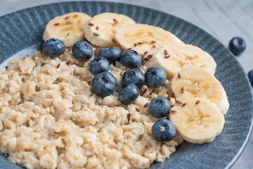 Close up view of healthy oatmeal porridge made of oats decorated with ripe blueberries, sliced banana and flax seeds served on blue gray plate on concrete background for breakfast in the morning