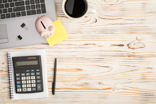 Top view photo of piggy bank binder clips yellow sticker note on laptop cup of drink pen and calculator on notepad on isolated bright wooden table background with copyspace