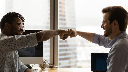 Diverse male colleagues giving fist bump at workplace. African guy greeting Caucasian mate, symbol of business partnership, racial equality, friendship at work. Respect, support, collaboration concept - obrazy, fototapety, plakaty