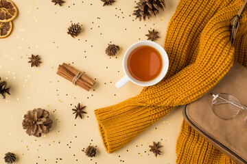 Fototapeta na wymiar Top view photo of yellow knitted pullover leather purse cup of tea glasses pine cones anise dried lemon slices cinnamon sticks and golden sequins on isolated light beige background