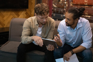 African ethnicity and Caucasian multiethnic male teammates staff members sit on sofa in office use digital tablet discuss collaborative project, share creative idea, consider new app. Teamwork concept